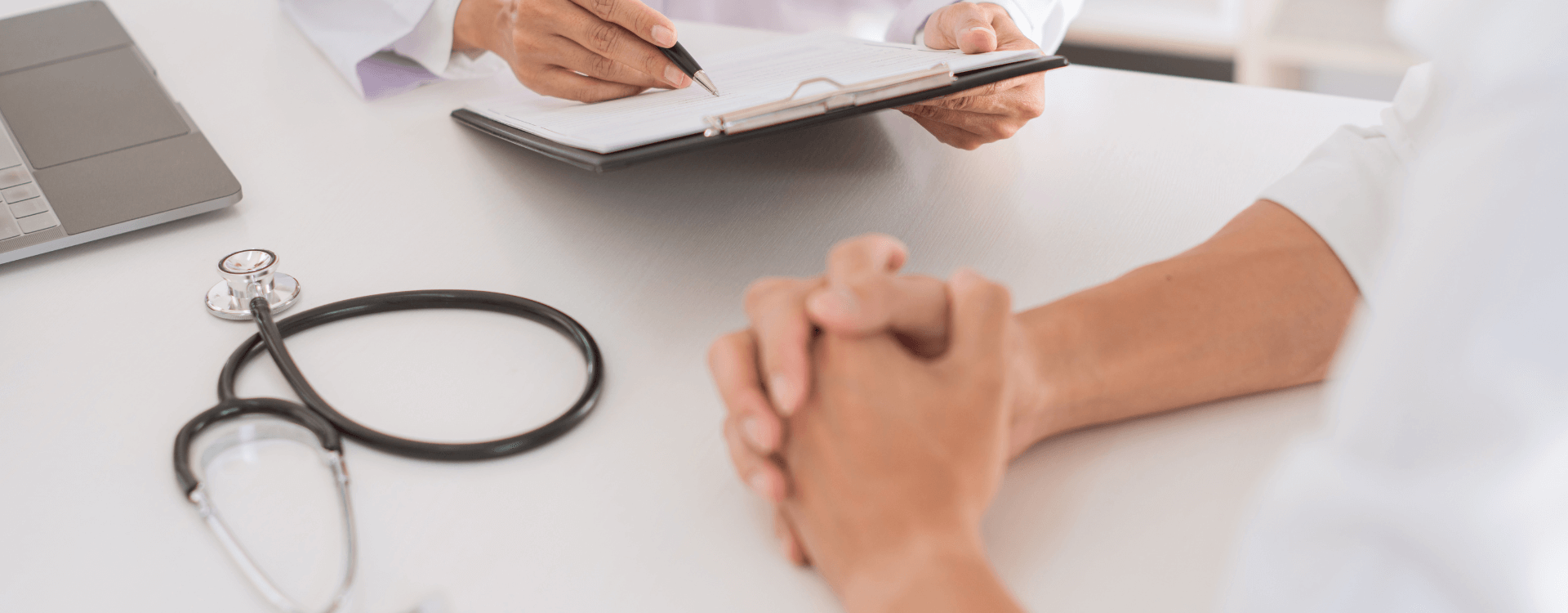 A doctor makes notes on a clipboard across a desk from a patient during a consultation